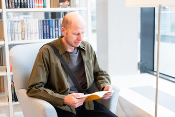 A bald man in his 40s wearing a green shirt reads a book in a library. A freelancer takes a break among the bookshelves.