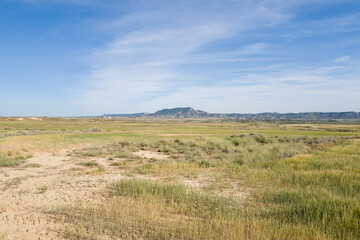 Fototapeta premium Wide stretches of green grass and dry earth extend toward low, rugged hills beneath a soft blue sky in Bardenas Reales, Spain. The landscape feels expansive, sunlit, and serene with natural textures