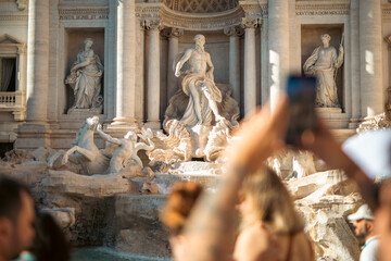 Tourists admire the famous Trevi Fountain on a sunny day in Rome, capturing memories with their smartphones.