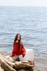 A young woman with long hair and blue eyes, in a white skirt and a terracotta sweater, sits on a tree stump against the backdrop of the sea