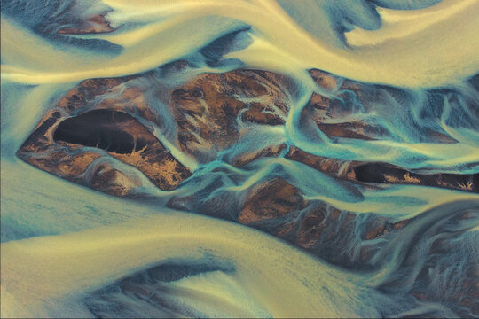 Aerial view of glacial river patterns create an ethereal landscape of swirling turquoise and gold, carving through the stark earth, Selfoss, Southern Region, Iceland.