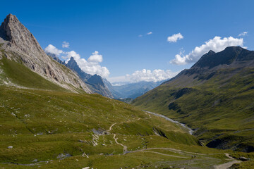 Obraz premium A narrow hiking path meanders through a wide green valley, flanked by steep rocky peaks and rolling grassy slopes under a bright blue sky. The landscape is sunlit and expansive, with dramatic mountain