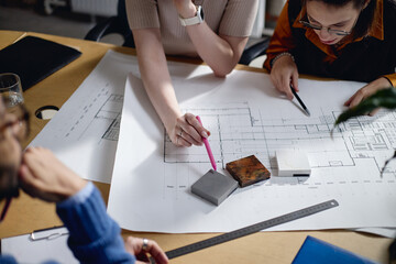 Two young adult women and multiethnic man collaborating on architectural project, reviewing blueprints and discussing material samples at office table