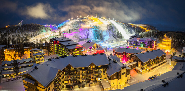 Aerial view of snow-laden rooftops and vibrant, illuminated ski slopes create a magical winter wonderland scene at night, Ruka, Lapland, Finland.