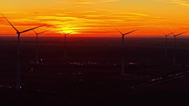 Aerial view of motionless wind farm turbines silhouetted against dramatic orange Latvia sunset sky over dark rural landscape on a windless day