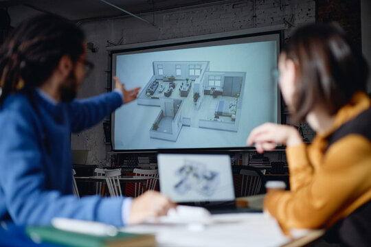 Caucasian man and Asian woman discussing architectural floor plan projected on screen in office setting, both sitting at table with laptop, focusing on building design presentation
