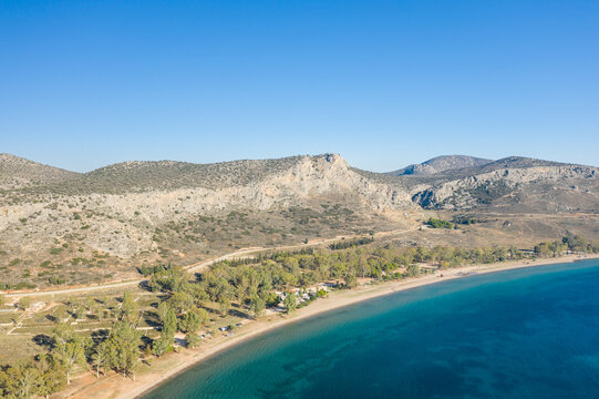 Aerial perspective of Karathona Beach near Nafplio, Greece, showing a quiet sandy shoreline bordered by clear blue water and rugged, sunlit hills with sparse vegetation under a cloudless sky.