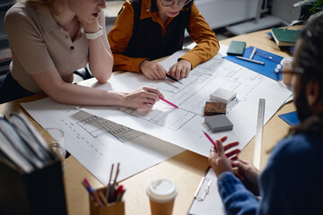 Diverse group of three young adult colleagues discussing architectural plans at desk, gesturing toward blueprints with pens, examining building material samples, collaborating on project