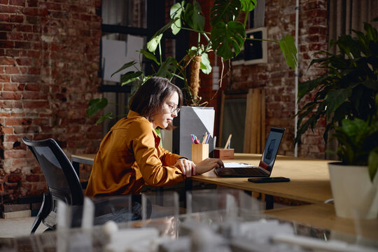 Young adult woman sitting at desk working on laptop in modern office space, concentrating on screen, surrounded by plants and office supplies, exposed brick wall background