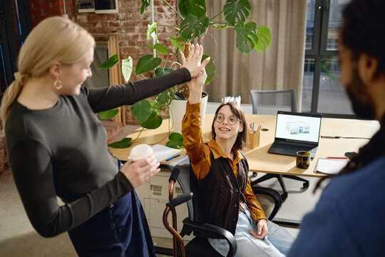Young Caucasian woman with disability sitting in wheelchair giving high five to smiling young Caucasian woman while holding coffee cup, multiethnic colleague standing nearby in office