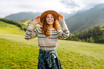 Happy woman in hat enjoys green hills at sunset, feeling free. A woman with backpack walks along a...
