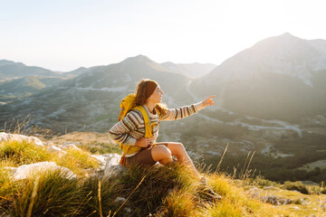 A young woman with hiking backpack stands on mountain trail on sunny day. Female hiker with a yellow backpack enjoys mountain scenery at sunset. Concepts of adventure, freedom. Active lifestyle. © maxbelchenko