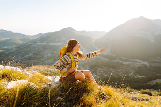A young woman with hiking backpack stands on mountain trail on sunny day. Female hiker with a yellow backpack enjoys mountain scenery at sunset. Concepts of adventure, freedom. Active lifestyle.