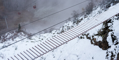 Rope bridge covered in snow across mountain gorge with cable car cabins and misty horizon in winter alps. Adventure and journey concept