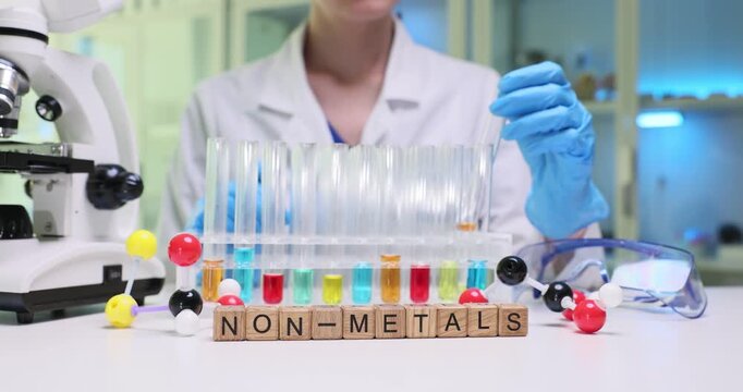 Text Non Metals on wooden blocks and scientist checking orange liquid leftovers in test tube. Female assistant works with color fluids in laboratory
