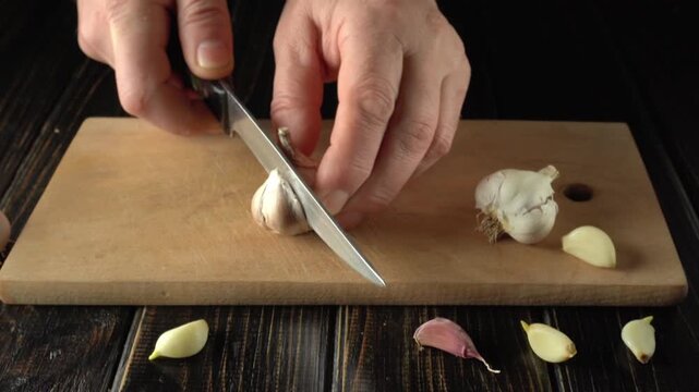 Hands slicing garlic bulb on wooden cutting board with garlic cloves scattered around