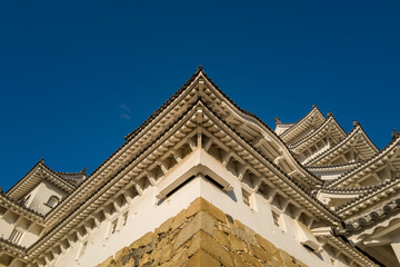 Obraz premium Striking upward angle of Himeji Castle's crisp white plaster walls and ornate tiled eaves meeting at a sharp corner, illuminated by warm sunlight against a deep blue sky.