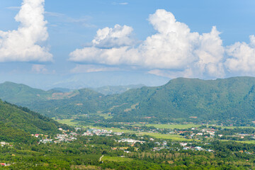 Naklejka premium A small town is nestled in a green valley surrounded by forested mountains and expansive rice fields under a bright blue sky with large, fluffy clouds. The landscape feels open, vibrant, and tranquil.