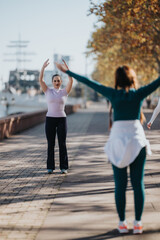 Obraz premium A woman stands facing the camera with arms raised while another person participates in a front-facing stretch along a riverside walkway.