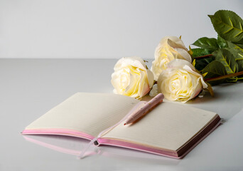 Pink diary with a pink ballpoint pen on white glass against the background of a bouquet of roses, space for text.