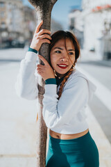A smiling woman in a white hoodie and teal leggings hugs a tree on a sunny city street. Casual athletic style and urban setting convey relaxed fitness and street fashion.