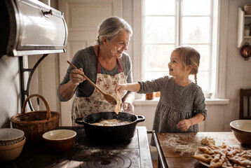 Happy senior grandmother and little granddaughter frying traditional angel wings pastry in deep oil on vintage stove during family cooking time in rustic kitchen
