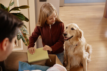 Girl unpacking box with golden retriever beside her at home