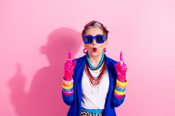 Aged glamorous mature woman in blue jacket and pink gloves poses playfully raised fingers confidence retro fashion against a bright pink studio background