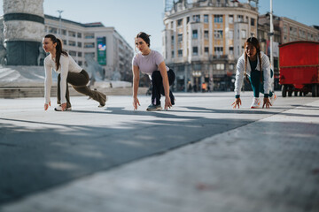 Obraz premium Three women in athletic wear crouch in a starting pose on a city plaza. The urban scene captures a dynamic fitness moment outdoors.