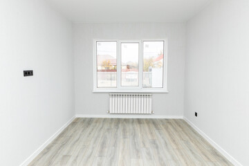 Obraz premium Empty interior room after renovations with white freshly painted walls and big window, allowing natural light, with a wooden floor, a radiator beneath the windows