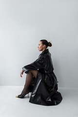 Naklejka premium Full-length portrait of a confident model in a glossy leather coat sitting in a creative pose. Edgy urban outfit featuring luxury footwear with gold accents and a sleek bun hairstyle.