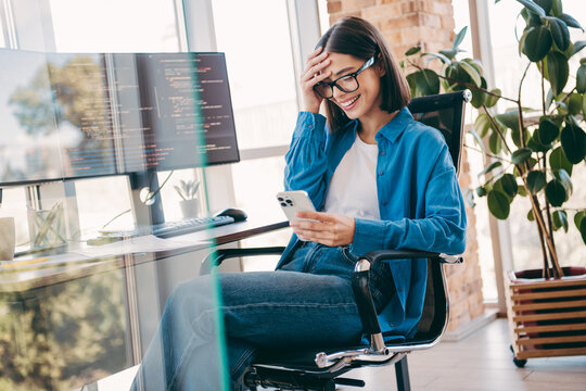 Female programmer in a bright loft office smiling at smartphone while coding on dual monitors in a modern tech business scene - Powered by Adobe