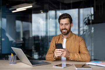 Smiling man concentrating on his smartphone while sitting at a modern office desk with a laptop,...