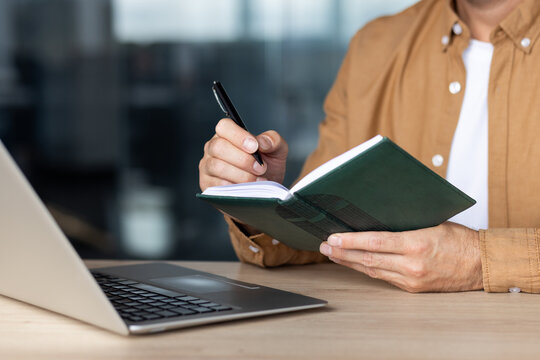 Man taking notes in a green diary, capturing ideas and planning tasks with a pen, sitting at a wooden desk with a laptop, focusing on organization and productivity