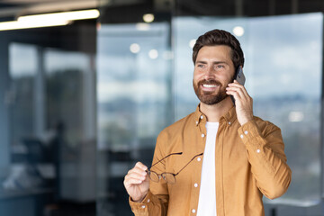Businessman talking happily on a mobile phone, holding his eyeglasses, while standing in a modern...