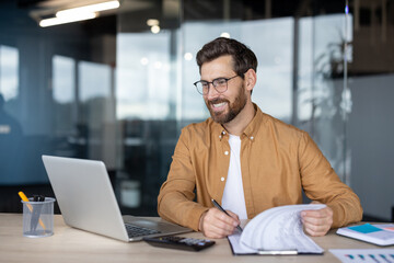 Smiling professional man wearing glasses concentrating on his work at a desk, reviewing documents while using a laptop and calculator in a contemporary office setting