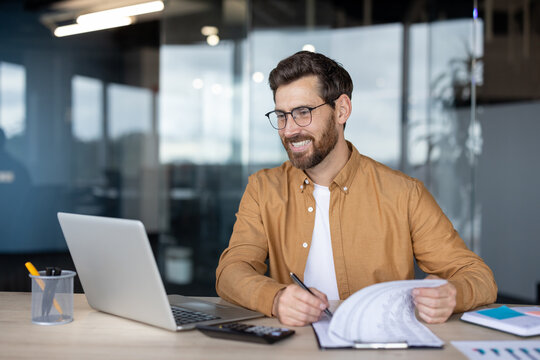 Smiling professional man wearing glasses concentrating on his work at a desk, reviewing documents while using a laptop and calculator in a contemporary office setting