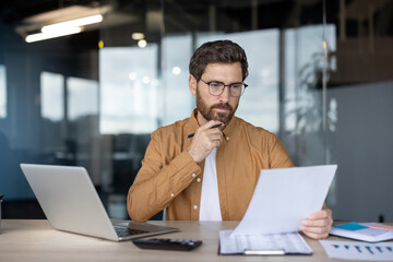 Thoughtful businessman in glasses analyzing financial documents and reports, holding a pen to his chin while working at a desk with a laptop in a modern office, focusing on strategy and planning