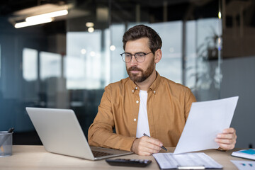 Bearded man wearing glasses processing data, reviewing financial reports, and working with documents and a laptop in a modern corporate office, focusing on productivity and planning