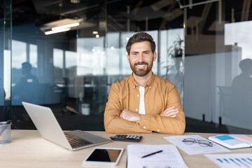 Young male entrepreneur sitting at office desk with laptop and documents, smiling broadly at camera, portraying success, startup, and professional achievement