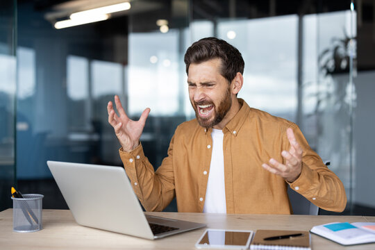Frustrated man experiencing work problems and stress while screaming at a laptop, showing anger and irritation with a difficult task or technical failure at the modern office desk