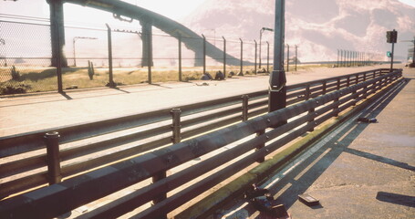 Sunlit empty bridge walkway at sunrise, long shadows from railings stretching across concrete pavement, fence and columns receding into distance, warm golden