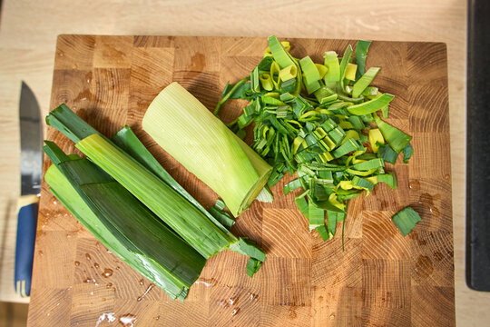 Chopping leeks on a wooden cutting board in a kitchen with a knife and fresh vegetables