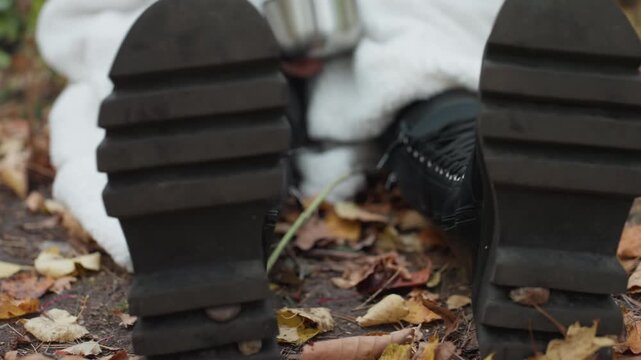 closeup leather boots on autumn leaves, woman wrapped in blanket holds metal cup, lowangle view highlights heavy tread and worn sole, gritty mood with earthy tones and soft overcast light