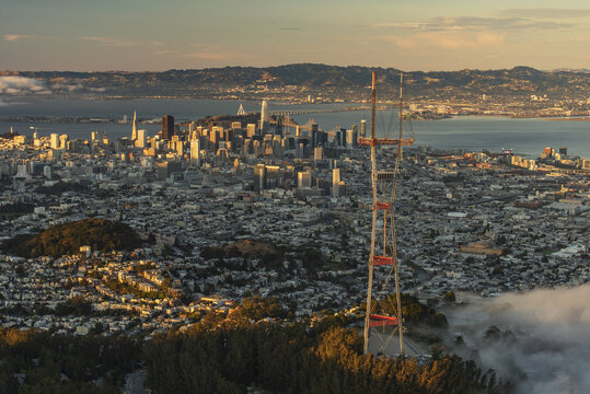 Aerial view of Sutro Tower piercing through the fog overlooking the sprawling cityscape and glimmering bay under a warm sunset glow, San Francisco, California, United States.
