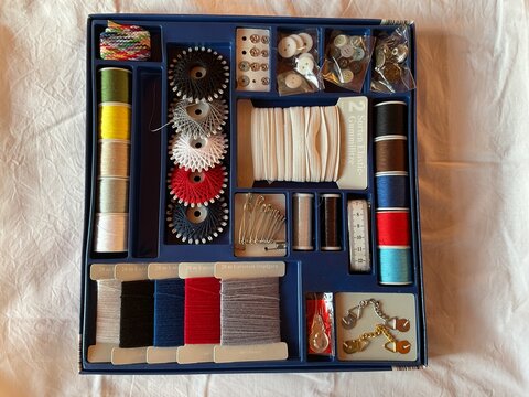 well-organized vintage-style sewing kit in a blue compartmentalized box, filled with colorful threads, various buttons, pins, safety pins, measuring tape, elastic, darning wool, and sewing accessories