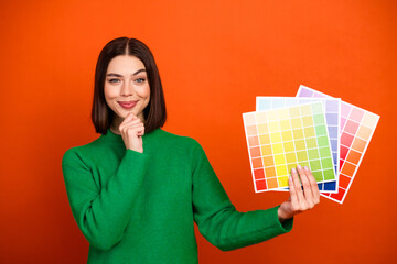 Young woman in a green sweater holding color swatches palette against an orange background to...