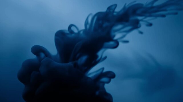 A captivating close-up of dark, opaque liquid expanding and dissolving gracefully into a deep indigo blue fluid. The organic shapes twist and swirl, creating a mesmerizing abstract display of fluid dy