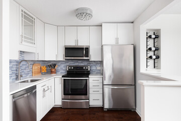 A white kitchen with stainless steel appliances, crystal light hanging from the popcorn ceiling, blue subway tile backsplash, and a wine rack mounted on the wall.