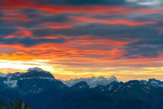 Aerial view of snow-capped peaks (Tofane and Sass Pordoi) pierce through the fiery canvas of a sunset sky, casting long shadows over the landscape, Trentino-South Tyrol, Italy.